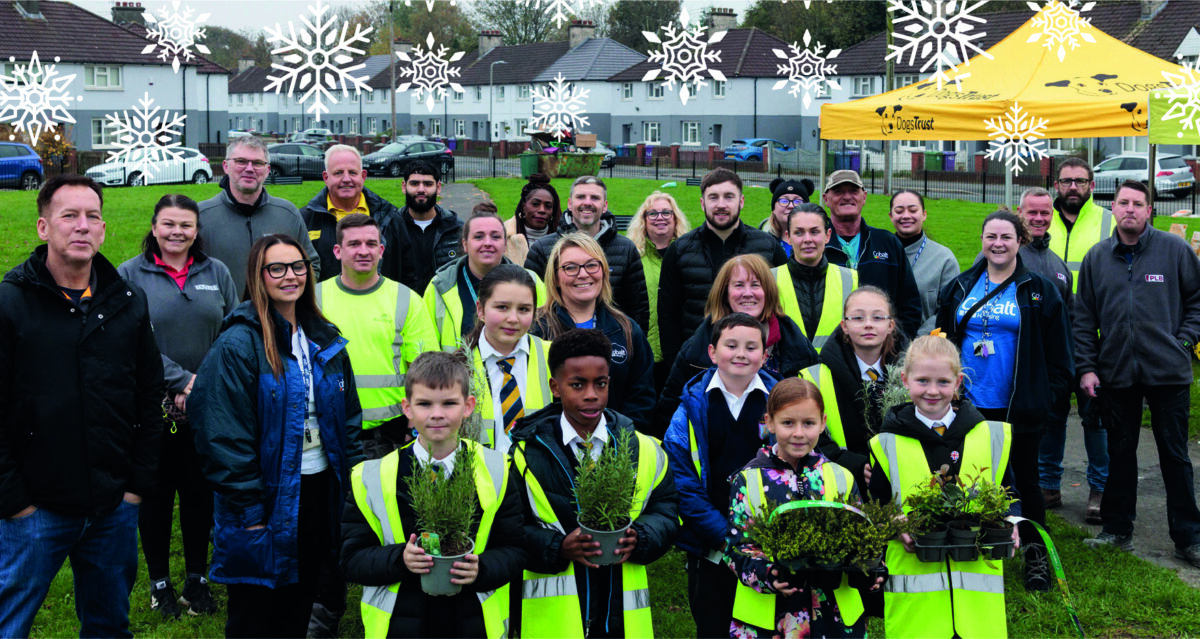 group of people of all ages outside attending a community event on grass, standing in a group posing for a photo in high vis jackets with children holding plants to be planted.