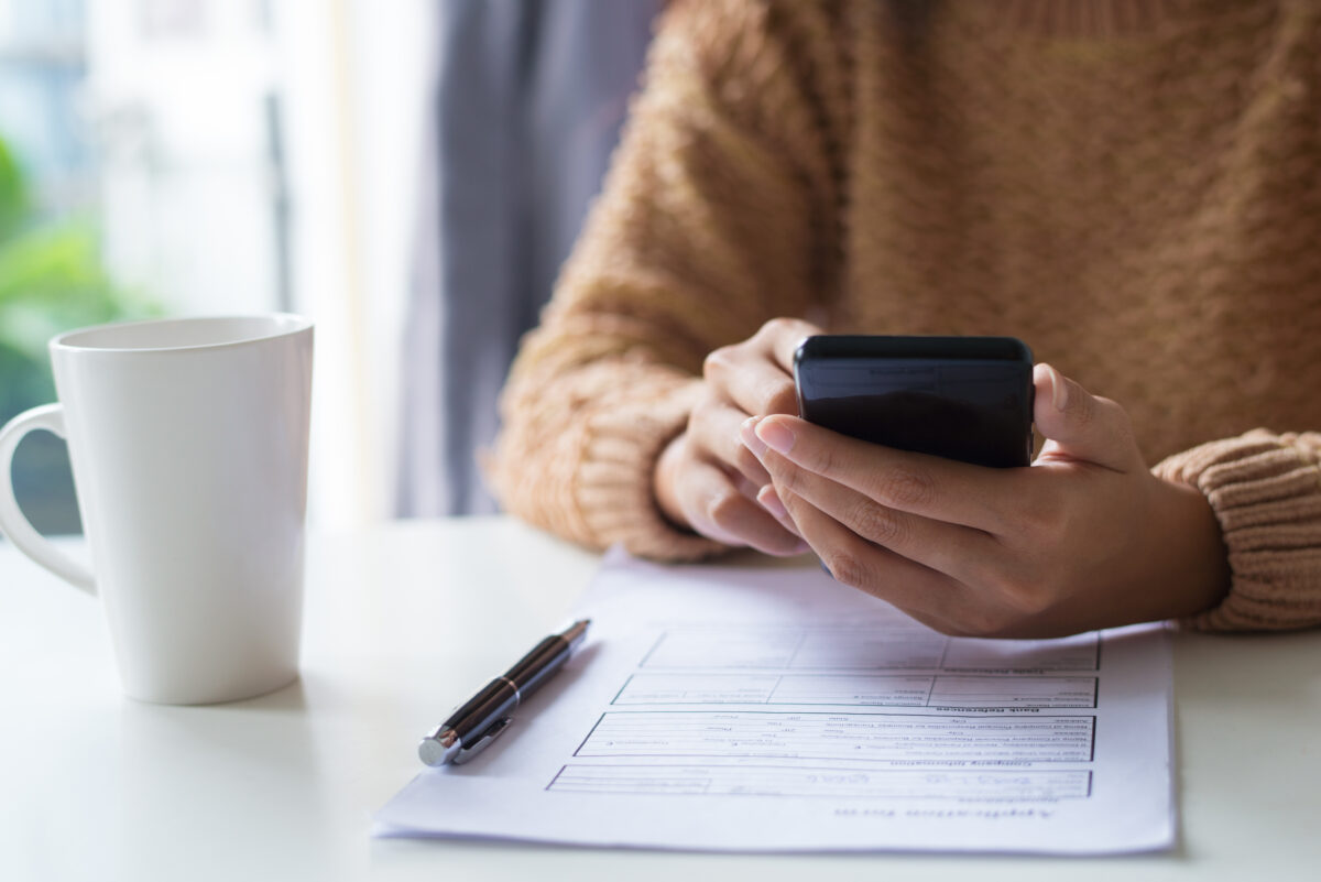Close-up of business lady using gadget while examining document. Unrecognizable woman in warm sweater sitting at table and checking message. Technology concept