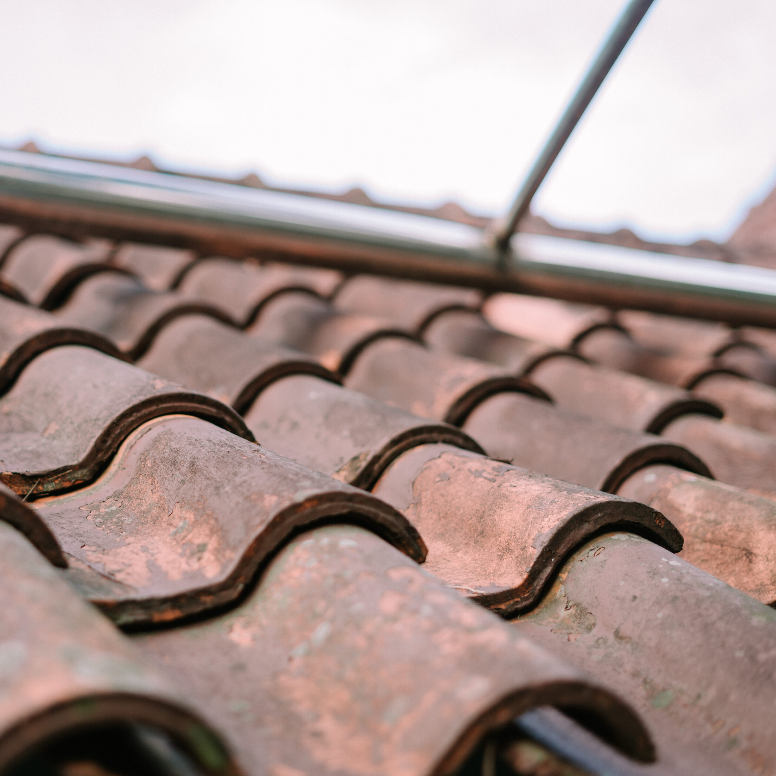 Close-up of rustic clay roof tiles in a wave pattern, with a metal pipe structure above. The weathered texture and mossy spots add character to the rooftop against a light sky.